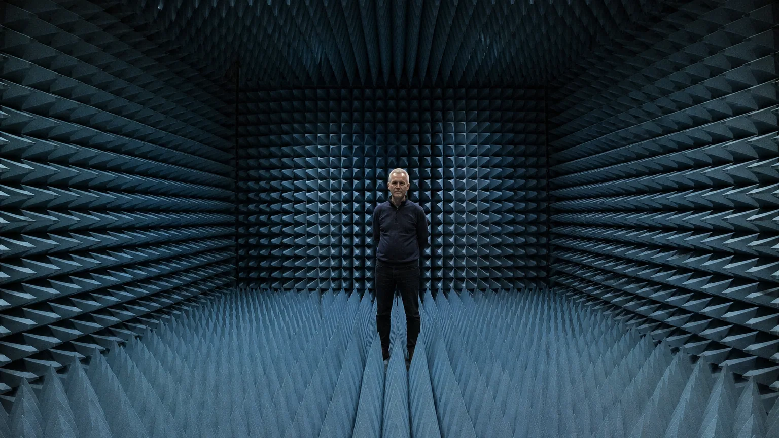 Intérieur d'une chambre anéchoïque tapissée de mousses acoustiques en pointes, personne debout au centre, lumière technique froide.