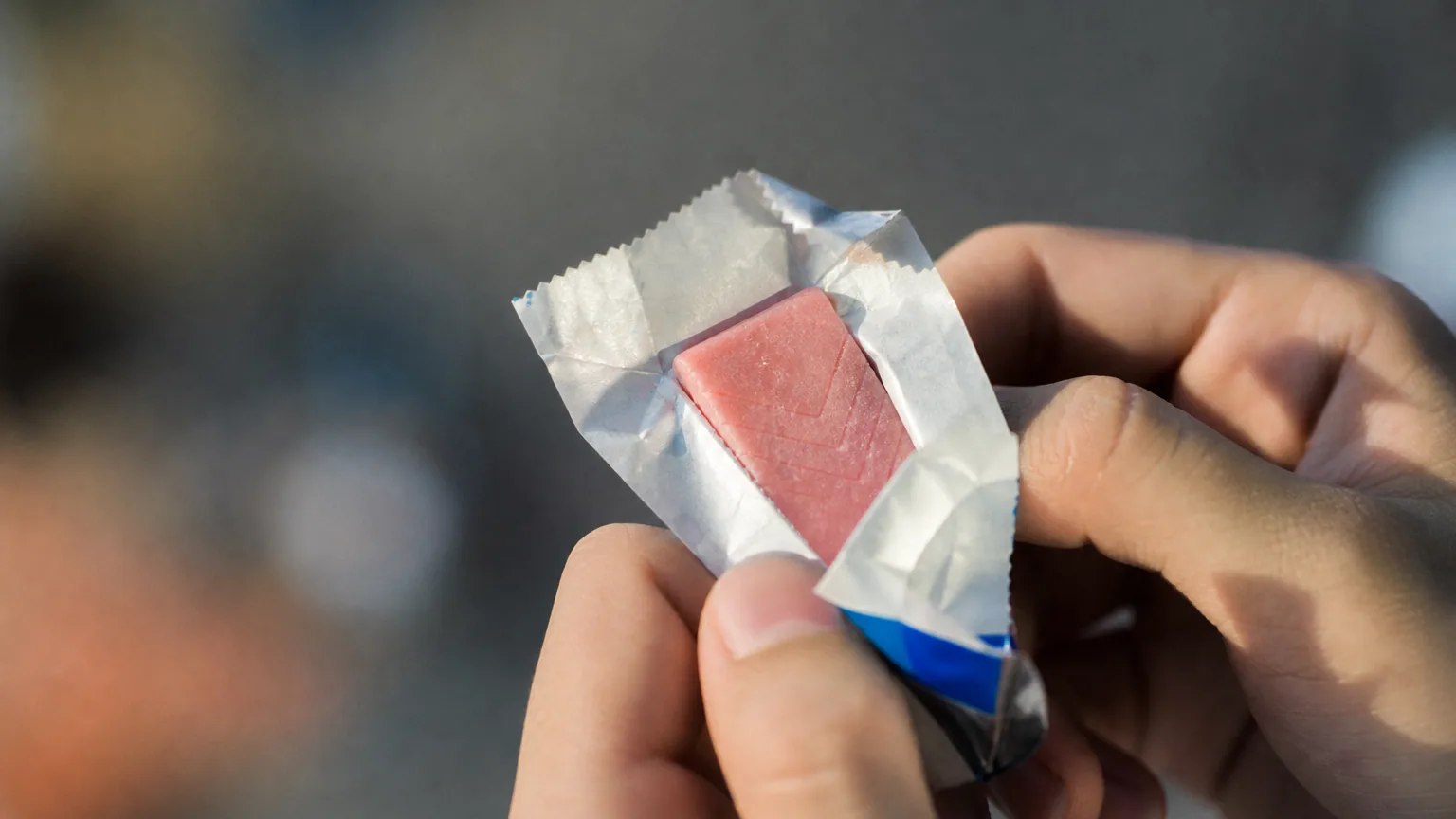 Main qui tient un chewing-gum rose en train d'être déballé, lumière naturelle douce, gros plan.