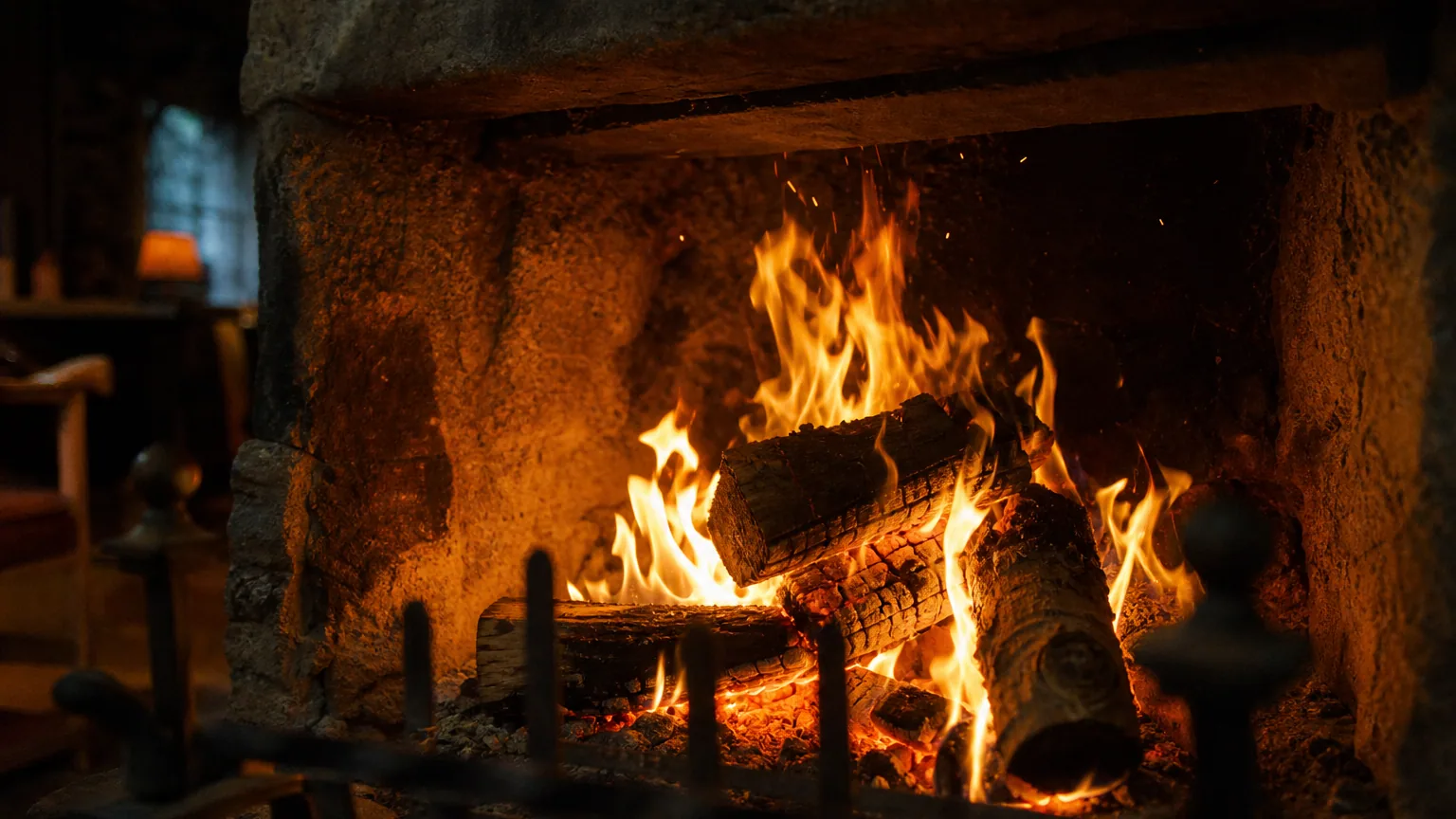 Flambée de bûches dans une cheminée en pierre, flammes orangées et quelques étincelles qui s'élèvent dans la pénombre d'un salon.