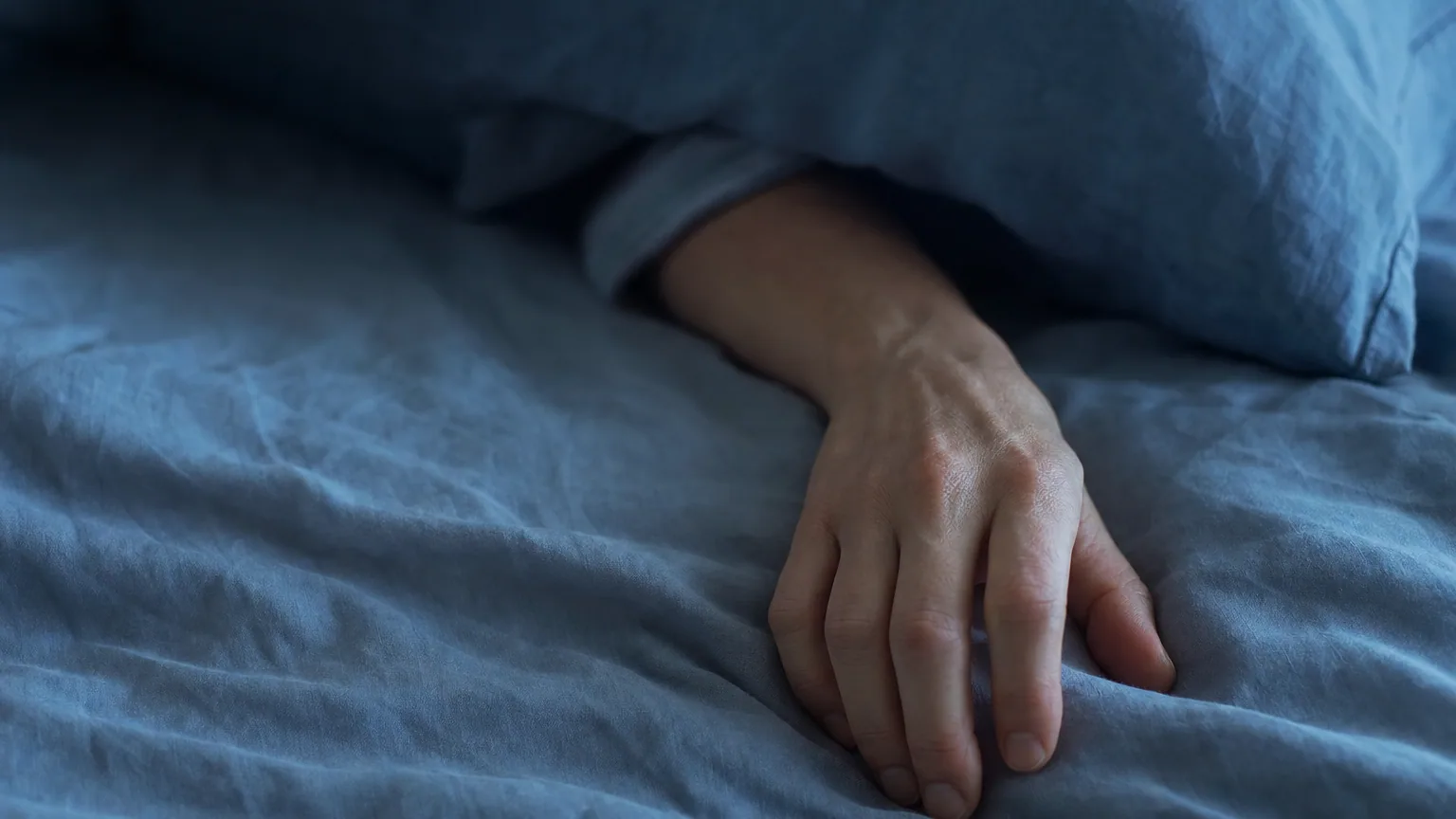 Une main d'adulte posée sur un drap froissé dans la lumière bleutée du petit matin, poignet replié sous l'oreiller.