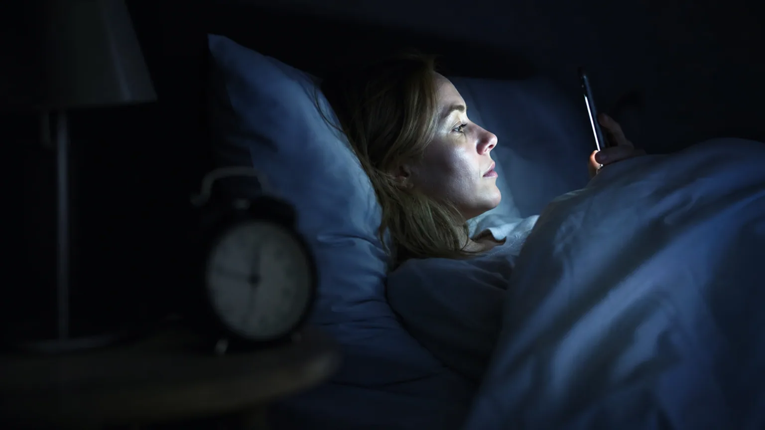 Visage éclairé par la lumière bleutée d'un smartphone dans une chambre plongée dans l'obscurité, la nuit.