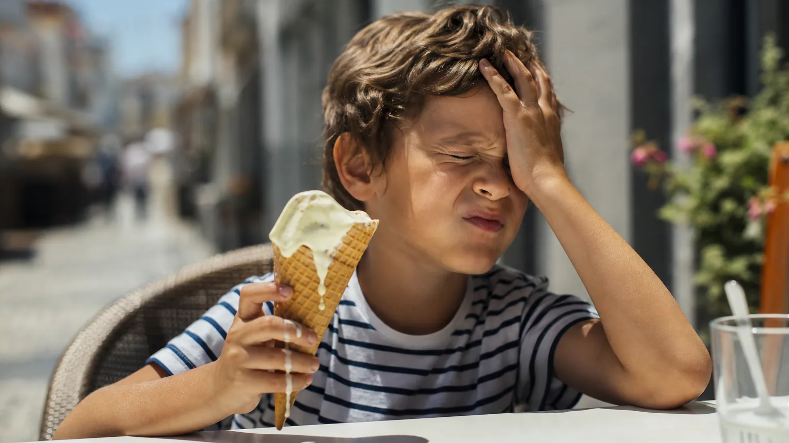 Un enfant grimace en pressant sa main sur son front, un cornet de glace à moitié fondu à la main, en plein soleil d'été.