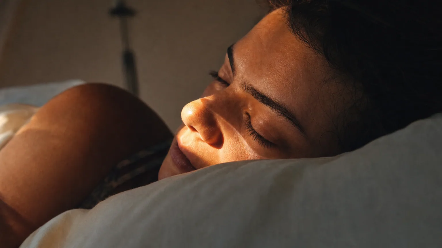 Femme allongée sur le côté dans un lit, lumière dorée soulignant son nez et ses narines au réveil.
