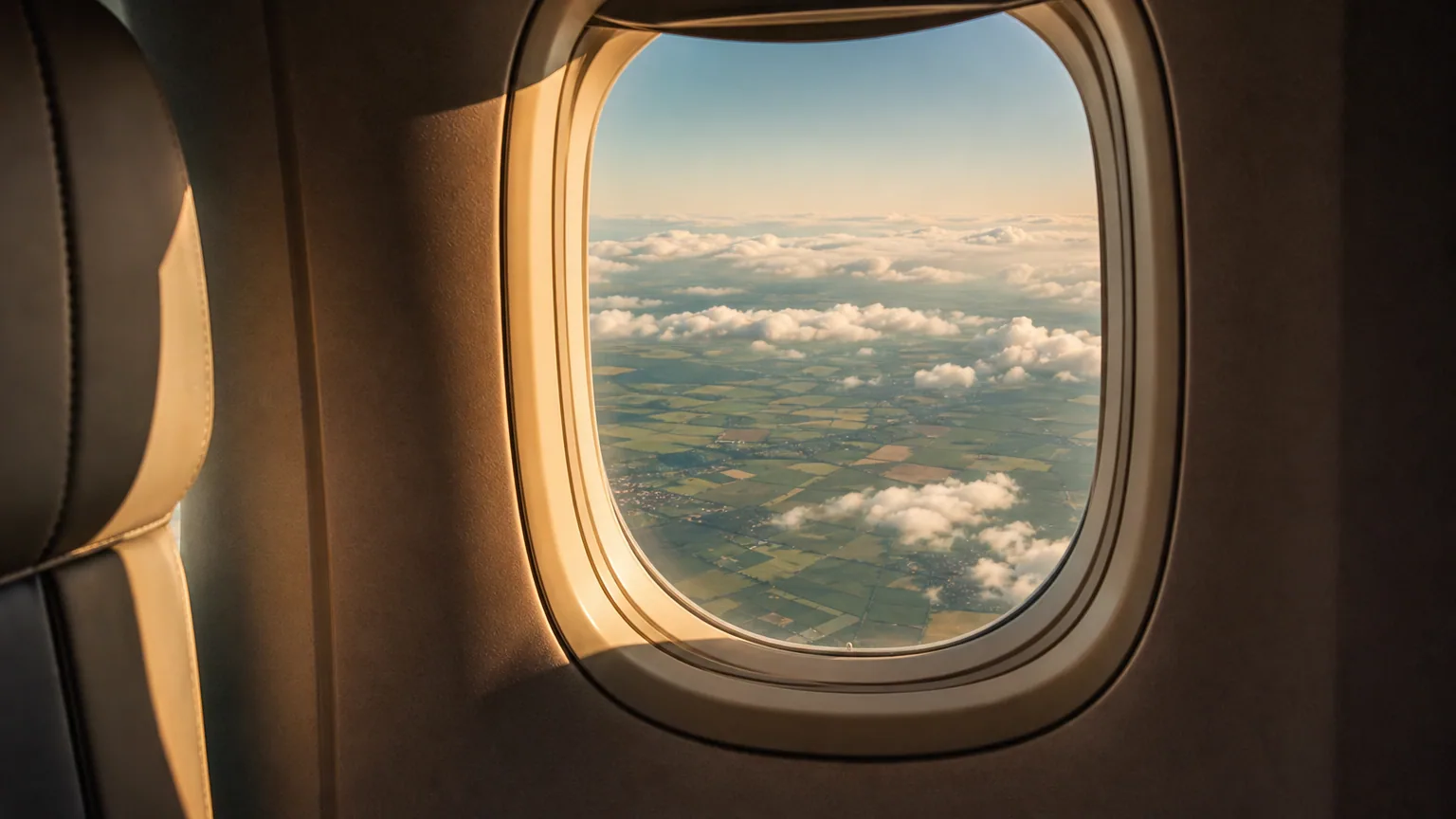 Vue rapprochée d'un hublot d'avion en phase de descente, le sol et les nuages en contrebas, la lumière dorée de fin de journée entrant dans la cabine.