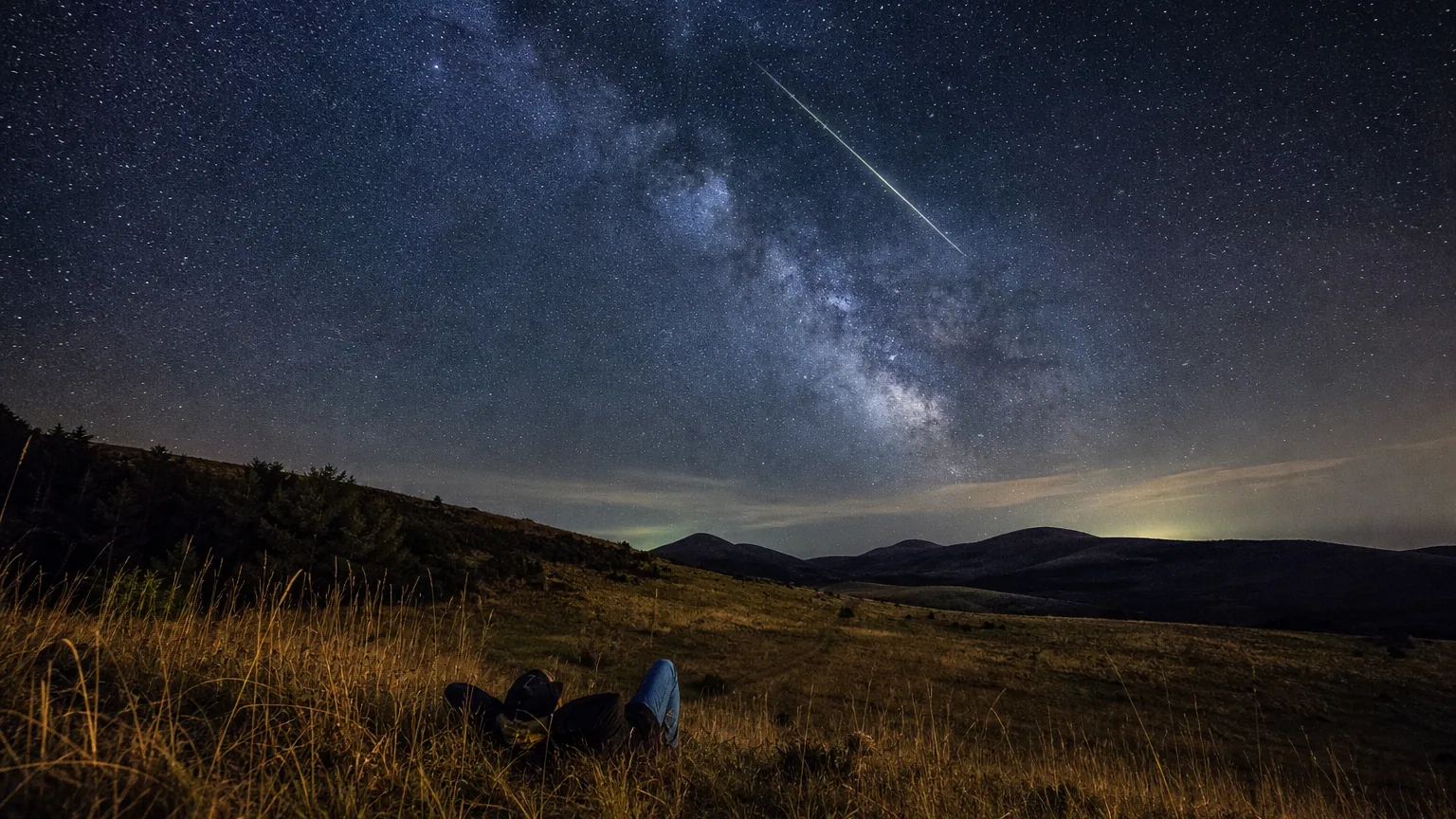 Un ciel nocturne d'été strié d'une longue étoile filante au-dessus d'une silhouette d'observateur allongé dans un champ.