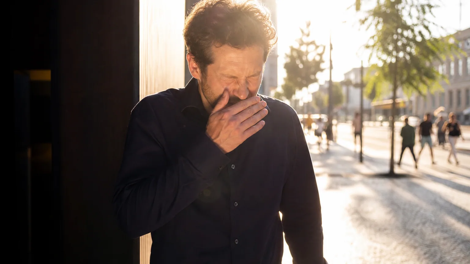 Homme qui plisse fortement les yeux et porte la main vers son nez en sortant à la lumière crue d'une journée ensoleillée, expression saisie sur le vif.