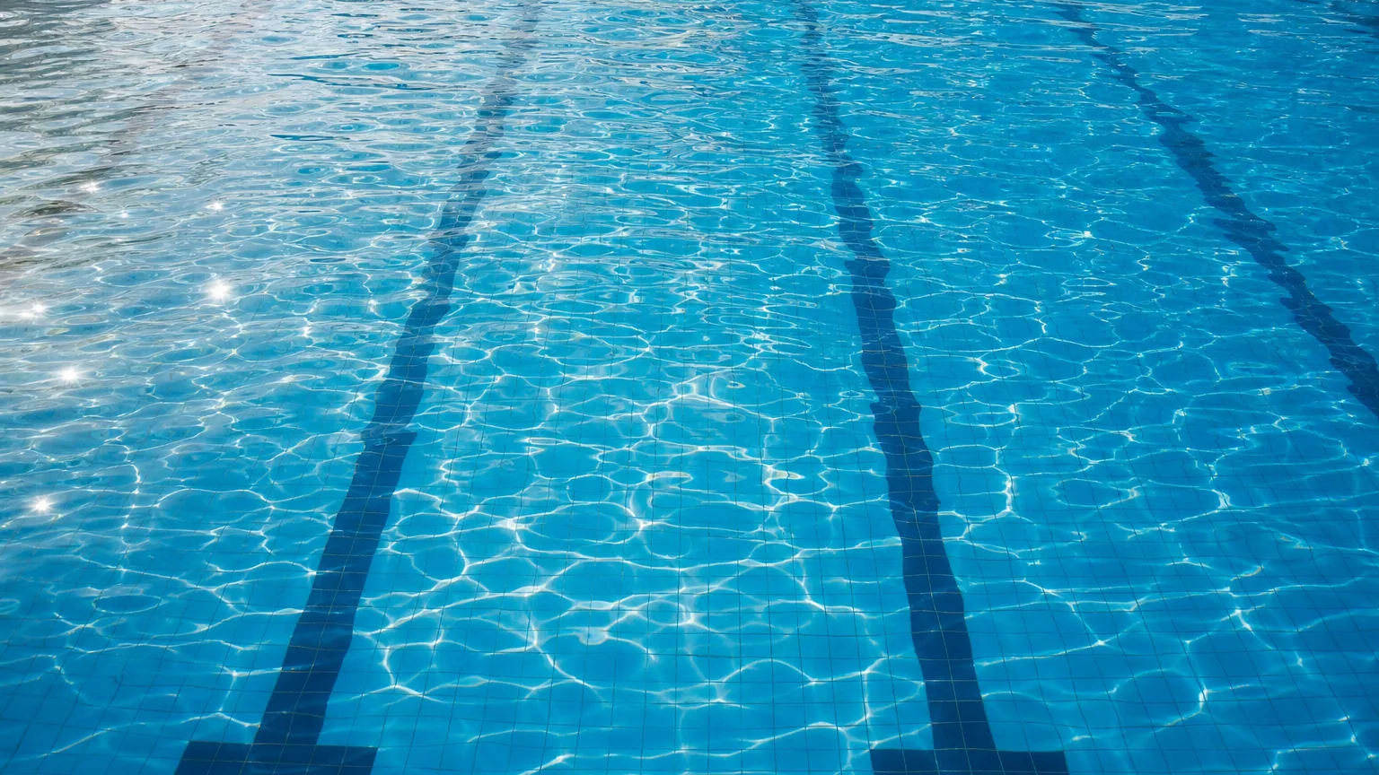 Vue plongeante sur le fond carrelé bleu d'une piscine sous un plein soleil d'été, la surface de l'eau parcourue de reflets et de lignes de lumière ondulantes.