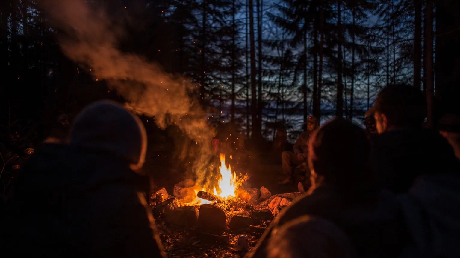 Un feu de camp le soir dans une forêt, flammes vives et épaisse fumée grise qui s'incline sur le côté, éclairant les silhouettes assises tout autour dans la pénombre.
