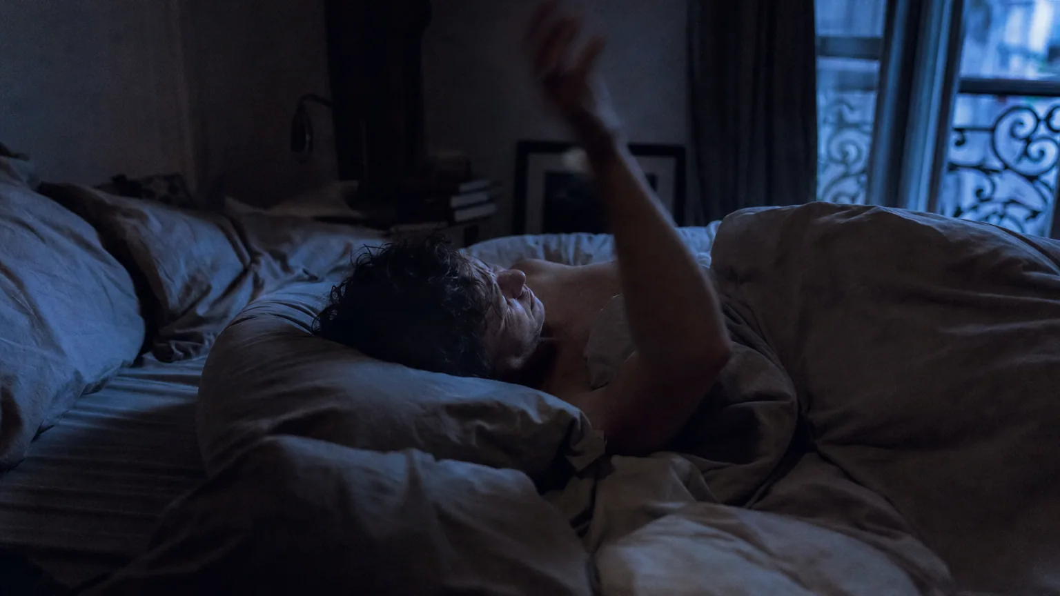 Une personne allongée dans un lit sombre dont le bras se soulève brusquement au moment de l'endormissement.