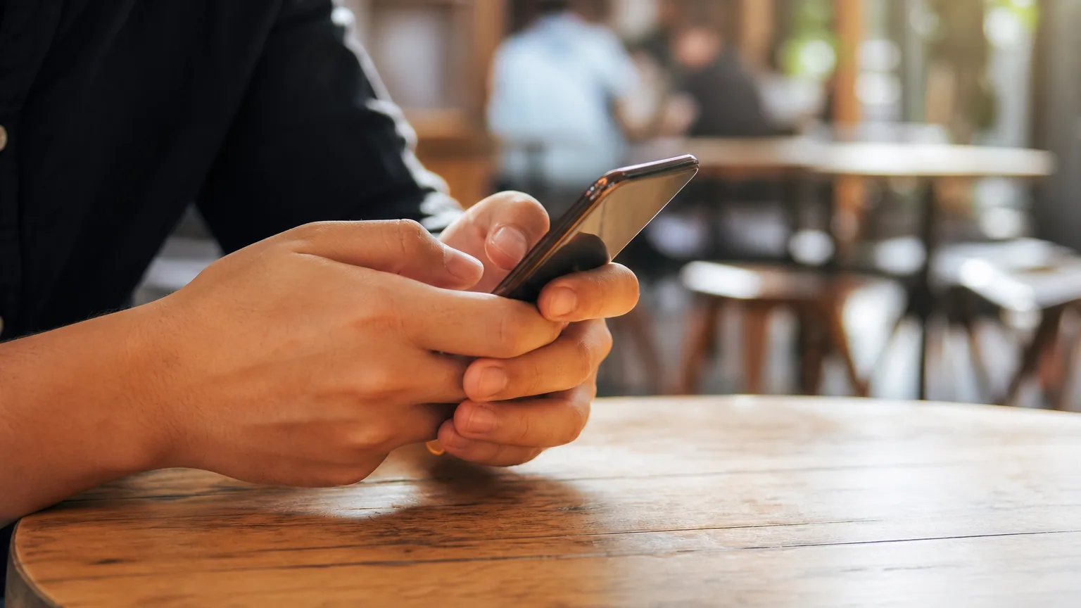 Gros plan d'une personne tenant un smartphone à deux mains dans un café lumineux, le pouce en approche de l'écran, arrière-plan chaleureux et flou.