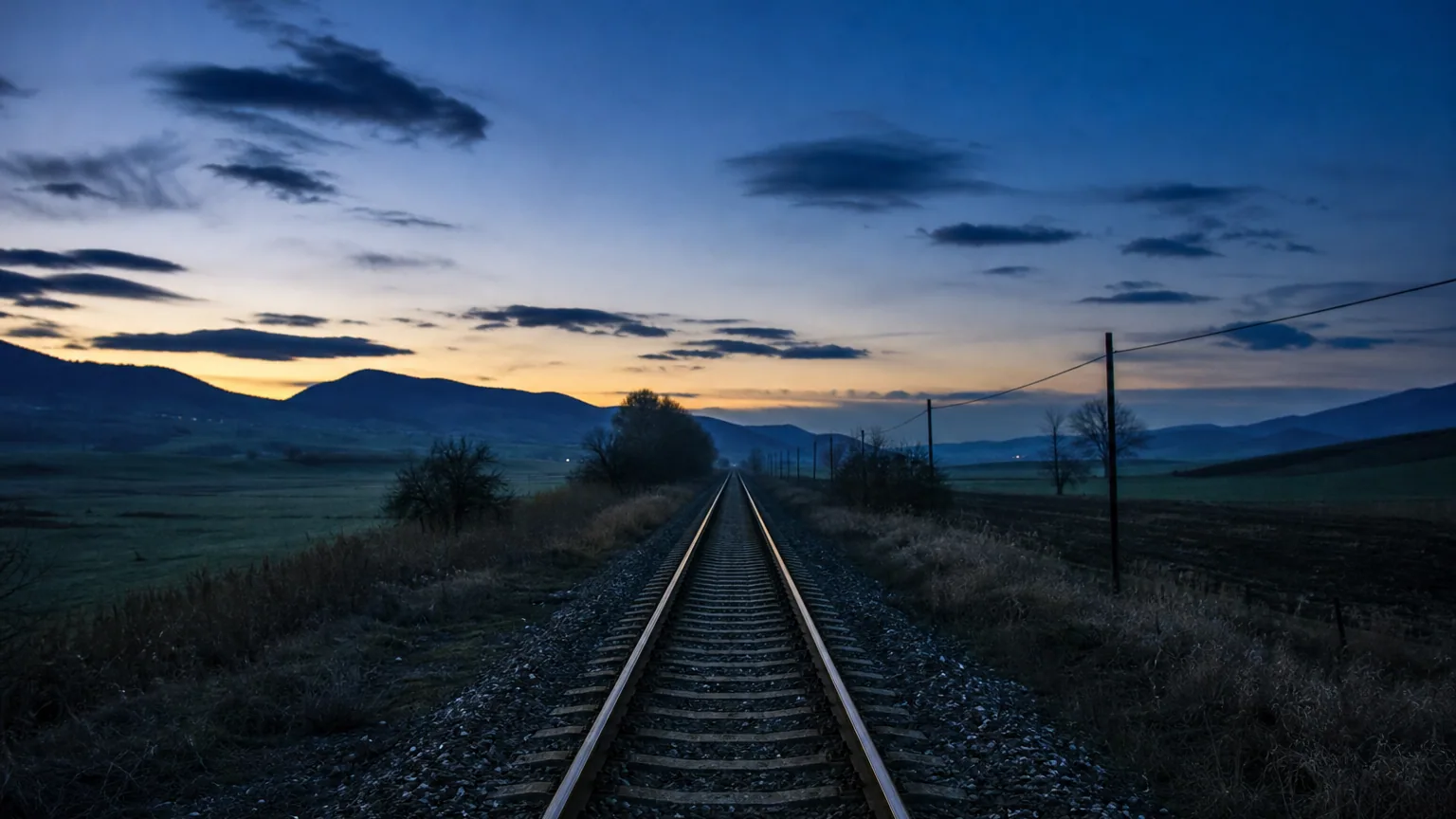 Une voie ferrée déserte qui file vers l'horizon dans la lumière bleutée du crépuscule, la campagne assoupie et silencieuse de part et d'autre des rails.