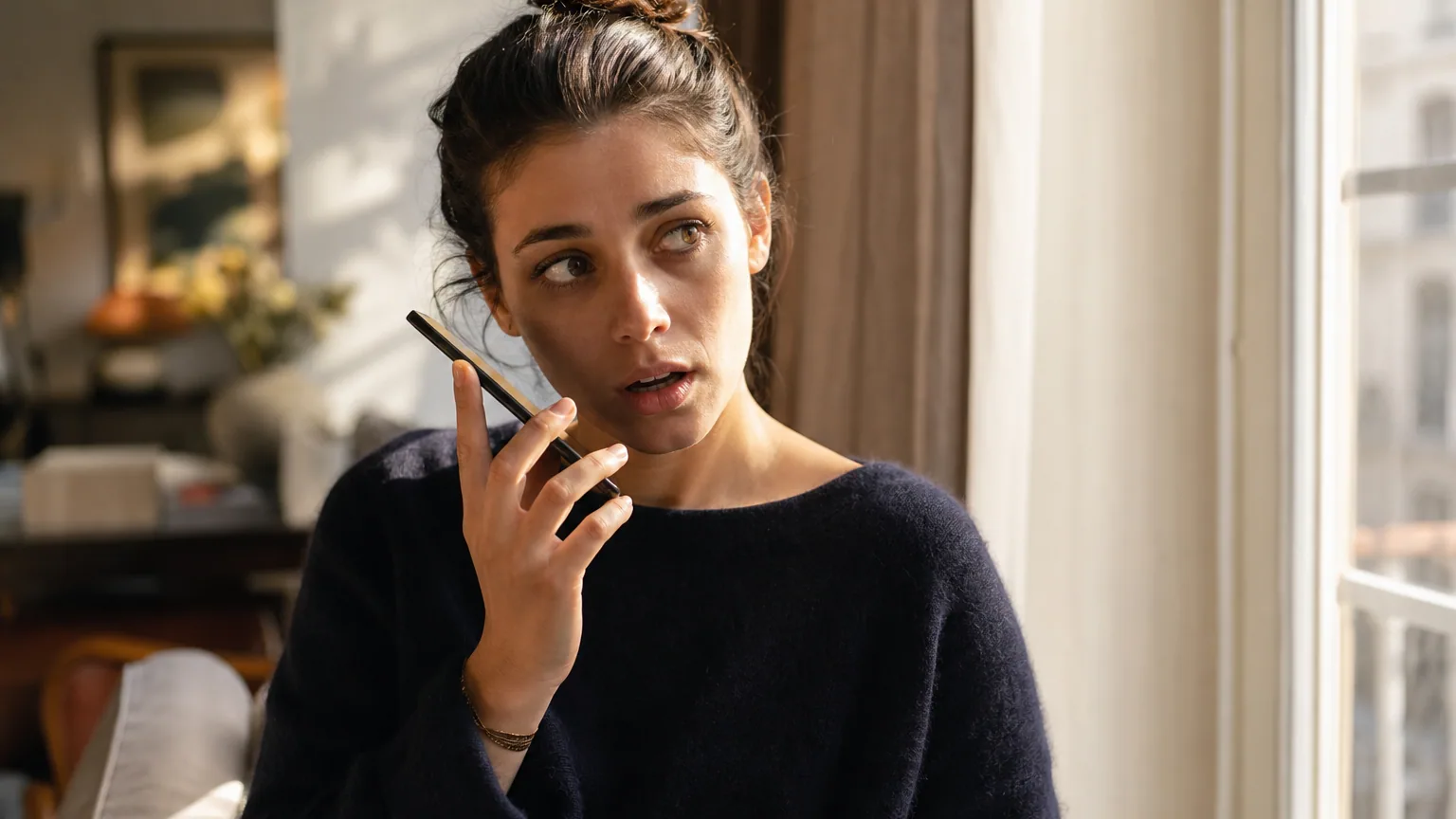 Une personne écoute un message vocal sur son smartphone, le visage légèrement crispé, dans la lumière douce d'un intérieur.