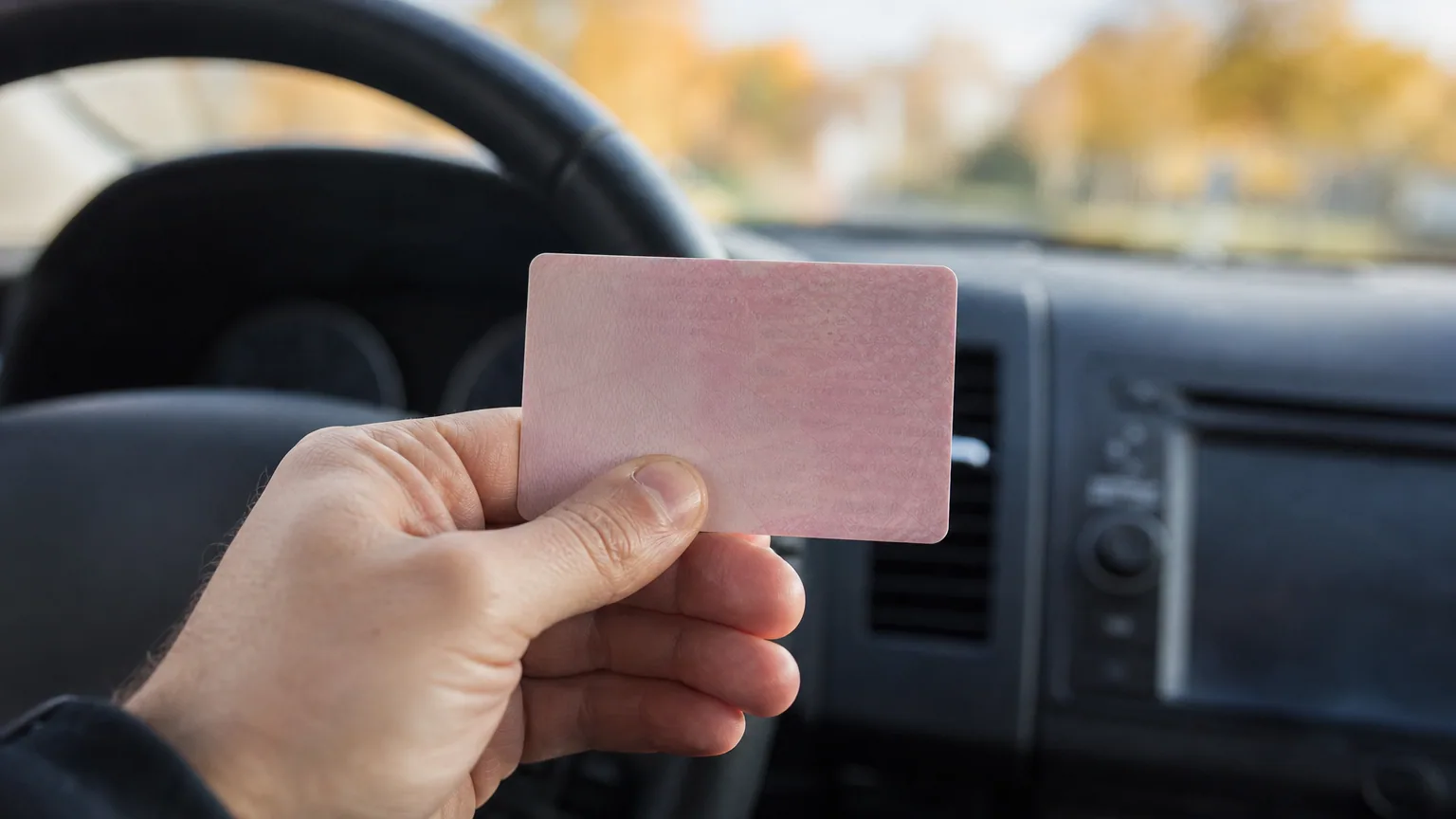 Conducteur tenant un permis de conduire rose au format carte bancaire au-dessus du volant de sa voiture garée.
