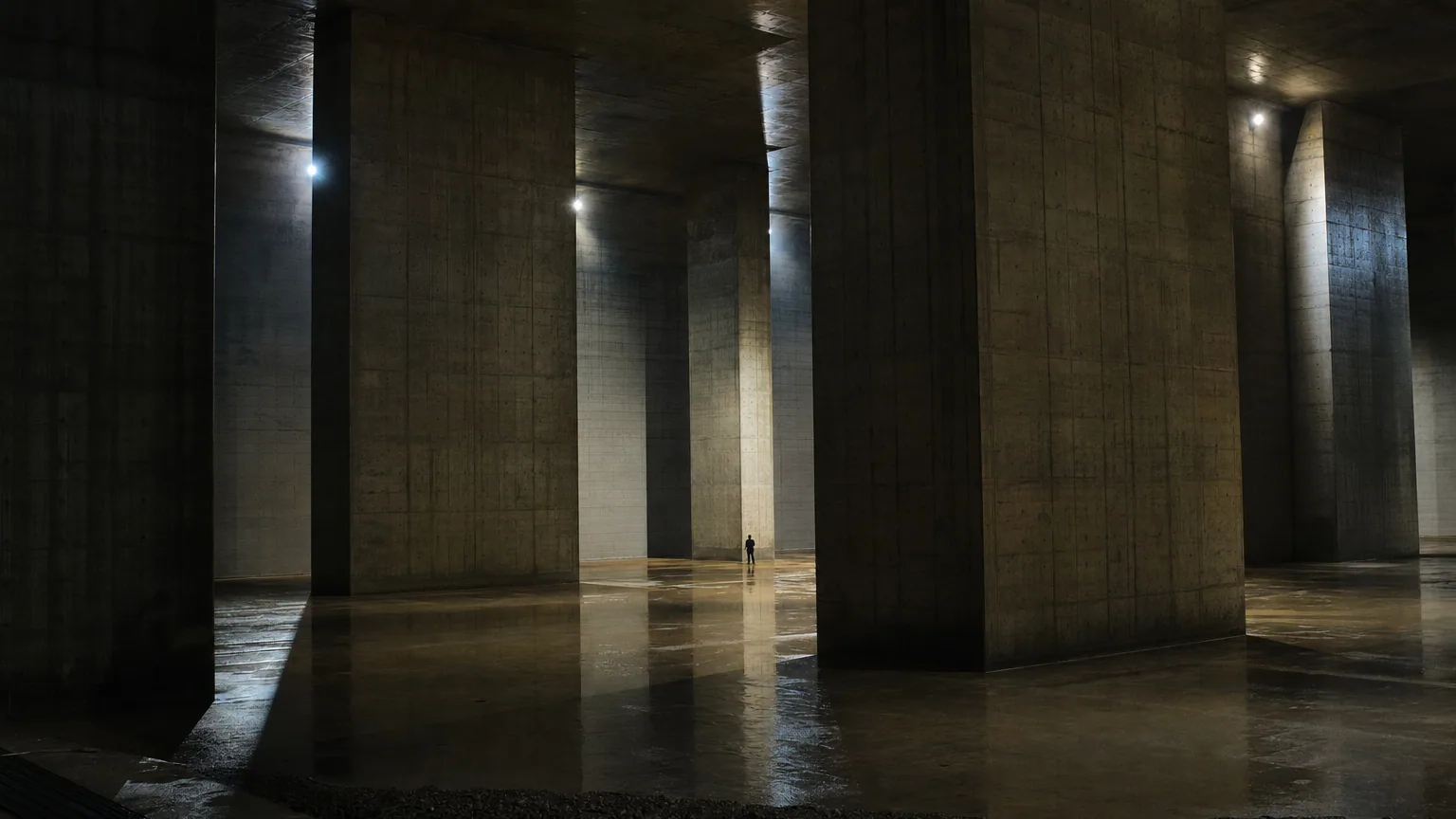 Salle souterraine en béton soutenue par d'énormes piliers, une personne minuscule au pied d'une colonne donne l'échelle.