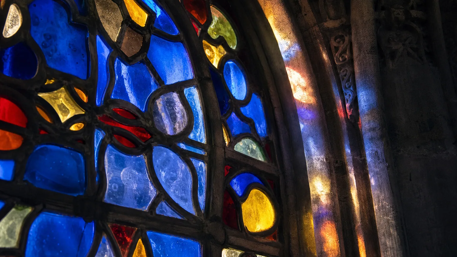 Détail d'un vitrail médiéval coloré traversé par la lumière, dont les pièces de verre montrent des épaisseurs et des bulles irrégulières.